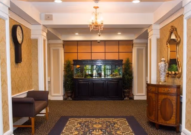 Interior hallway with patterned carpet and wallpaper, featuring a large fish tank centered against the far wall flanked by two potted plants. On the left side, there is a dark upholstered bench beneath a wall clock. On the right side, there is a wooden console table with a decorative vase and a gold-framed mirror above it. A chandelier hangs from the ceiling.