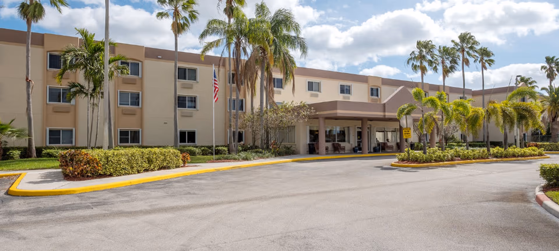 Exterior front view of a three-story senior living facility building with beige and brown walls, surrounded by palm trees and landscaped bushes. There is a covered entrance with seating and an American flag on a flagpole near the entrance. The sky is partly cloudy.
