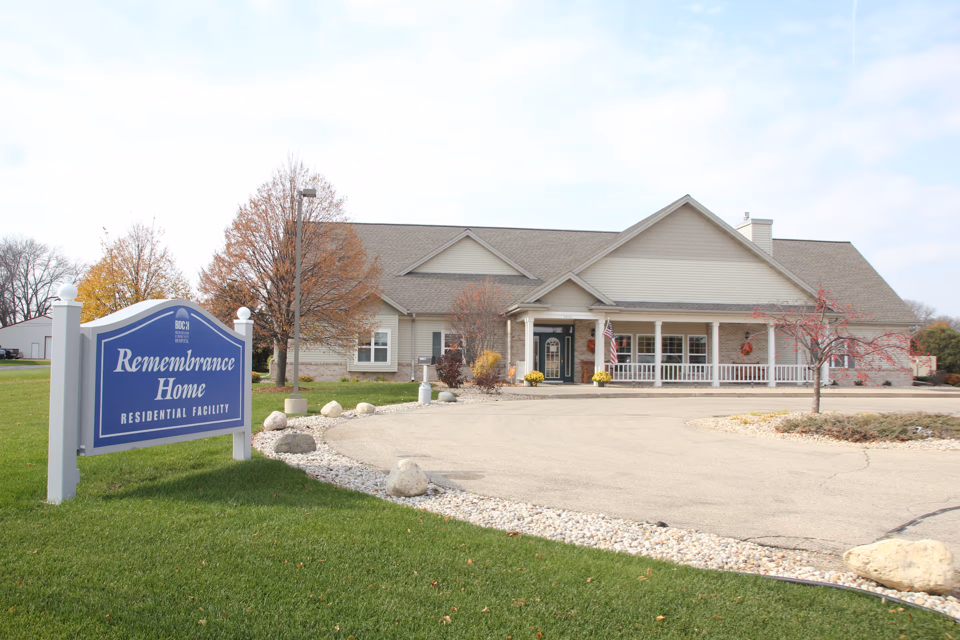 Front exterior of a one-story residential care building with a porch and a blue 'Remembrance Home' sign on the lawn.