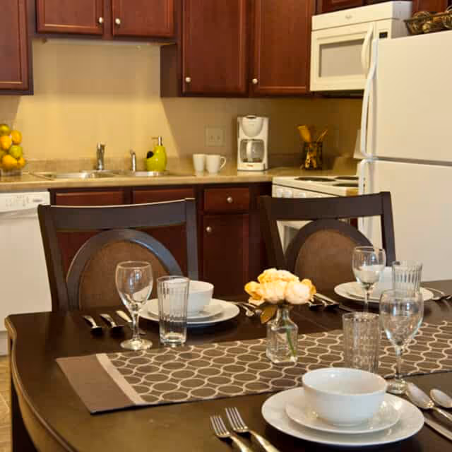 Dining table set with plates, bowls, glassware and a small flower vase in front of a kitchen with dark wood cabinets, white appliances, and a coffee maker.