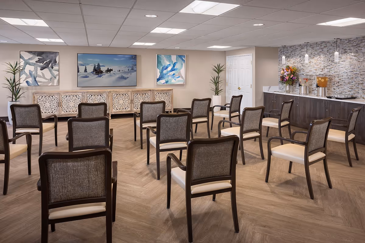 A well-lit room with multiple chairs arranged in rows facing a wall with three framed artworks. The room features a wooden floor, two tall potted plants, a decorative wooden cabinet, and a countertop with a mosaic tile backsplash, flowers, and beverage dispensers.