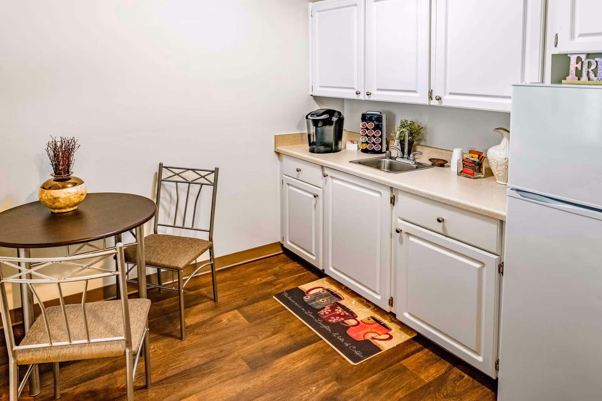 Small kitchen area with white cabinets, a countertop with a sink, a coffee maker, and a coffee pod holder. There is a white refrigerator on the right side. To the left, there is a small round dining table with two metal chairs with beige cushions. The floor is wooden, and there is a decorative mat in front of the sink.
