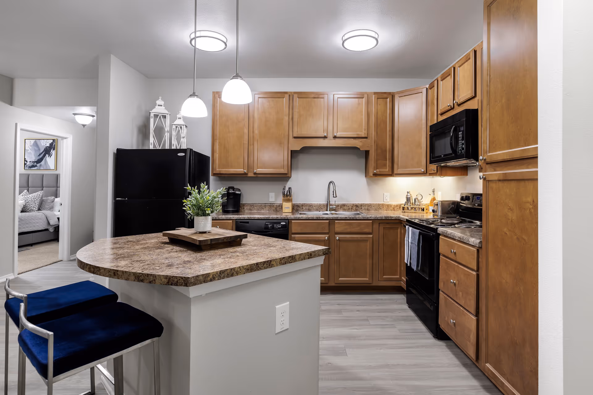 Modern kitchen with wooden cabinets, black appliances including a refrigerator, stove, microwave, and dishwasher. A countertop island with two blue cushioned stools and a small potted plant is in the foreground. Pendant lights hang above the island. A bedroom with a bed and artwork is visible through an open doorway to the left.