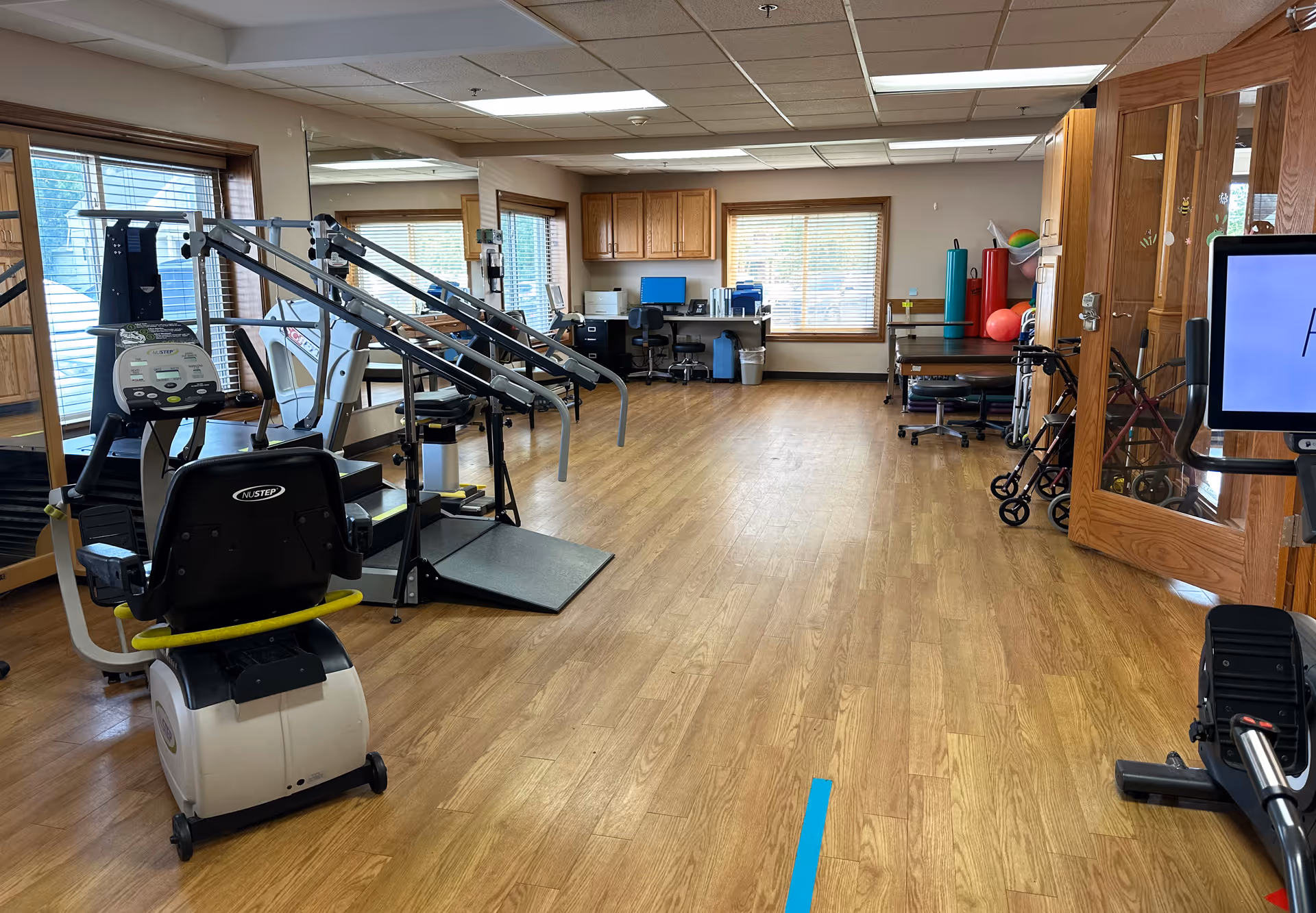 A spacious room with wooden flooring and large windows letting in natural light. The room contains exercise equipment including a recumbent bike, parallel bars for walking support, and other physical therapy tools. There is a desk with a computer and office chair against the far wall, and various therapy balls and equipment are stored on shelves. The room appears to be a rehabilitation or physical therapy area within a healthcare facility.