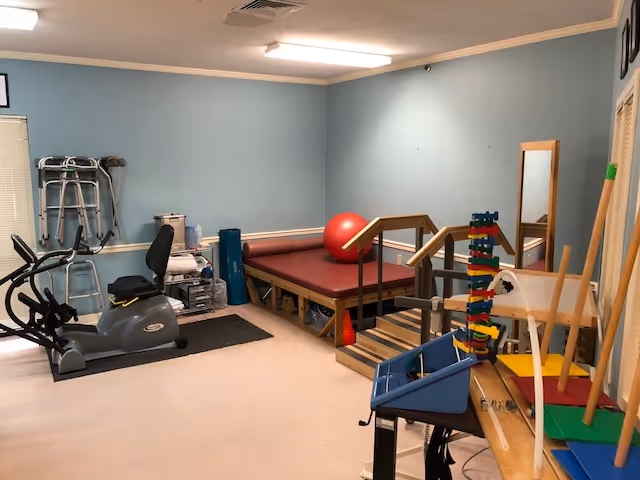 A therapy or rehabilitation room with exercise equipment including a stationary bike, a walker, a red exercise ball, padded therapy tables with handrails, and various therapy tools and supplies on shelves and tables against light blue walls.