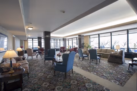 Spacious senior living lounge with upholstered chairs, sofas, side tables, lamps, and large windows along one wall.