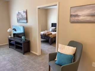 Small living area with an armchair and TV stand, wall artwork, and a bedroom visible through an open doorway.