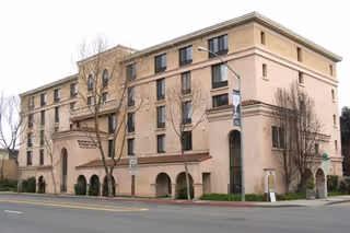 Exterior view of a multi-story beige building with arched windows and entrances, leafless trees in front, and a street with a speed limit sign in the foreground. The building has a sign that reads Marymount Villa.