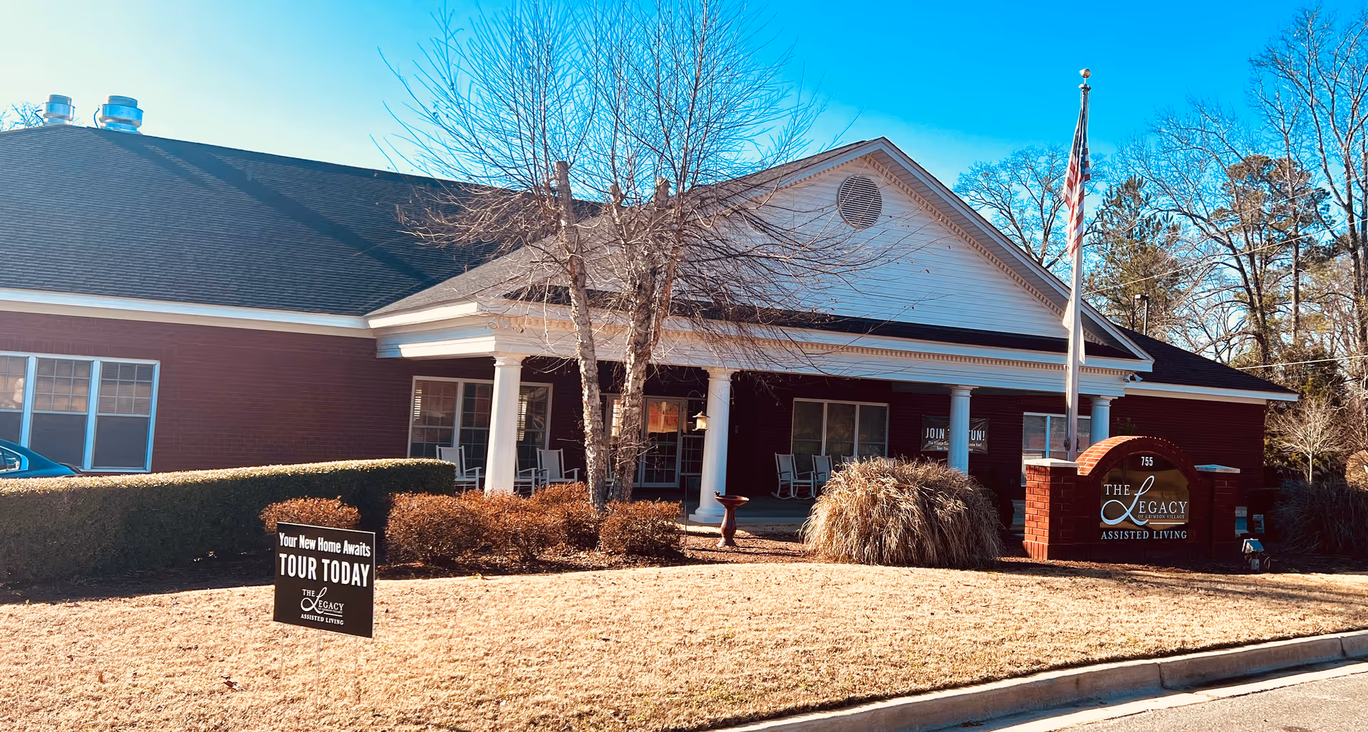 Exterior view of The Legacy of Crimson Village Assisted Living building with a brick facade, white columns, and a covered porch with rocking chairs. There is a sign on the lawn that says 'Your New Home Awaits TOUR TODAY' and another sign near the entrance displaying the facility name. The sky is clear and blue, and there are leafless trees and bushes around the building.