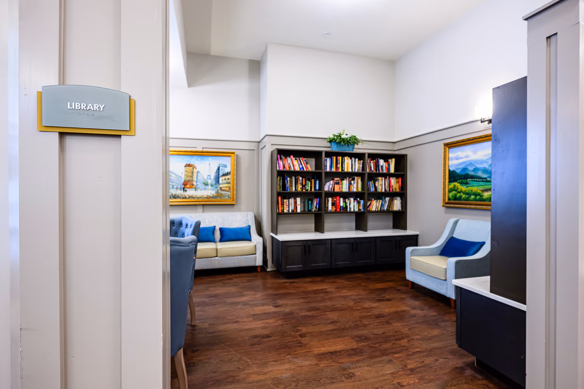 Interior view of a library room in a senior living facility with wooden flooring, a bookshelf filled with books, two paintings on the walls, and comfortable seating including a sofa and armchairs with blue cushions.