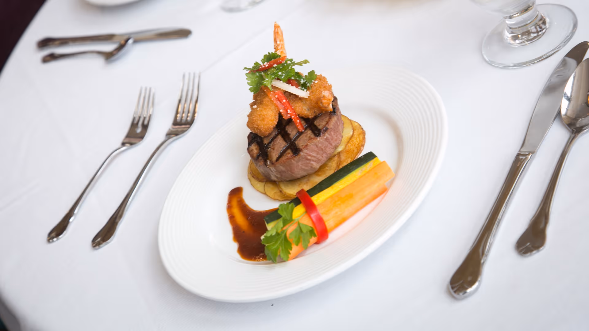 A gourmet plated meal featuring a grilled steak topped with fried shrimp and garnished with herbs, served on a bed of sliced potatoes with a side of colorful vegetable sticks and a drizzle of sauce on a white plate. The plate is set on a white tablecloth with silver cutlery and a glass of water nearby.