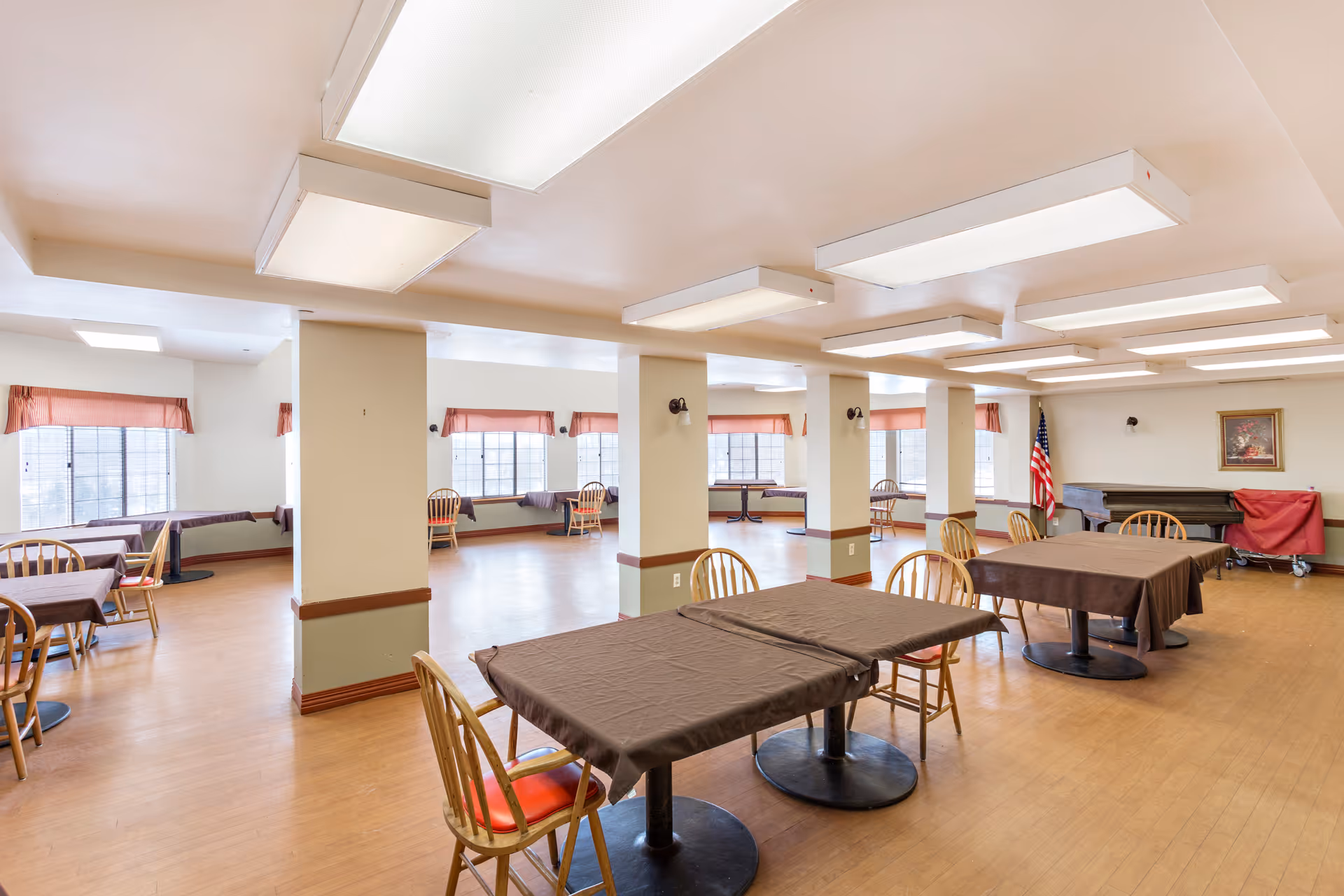 A spacious dining room with multiple tables covered in brown tablecloths and wooden chairs with red cushions. The room has large windows with red valances, several white columns, and bright overhead lighting. An American flag and a piano covered with a red cloth are visible in the background, along with a framed picture on the wall.