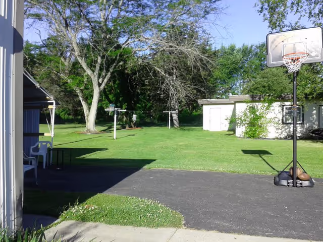 Outdoor area with a basketball hoop on a paved surface, green grass lawn, large trees, a white shed, and a covered patio with white chairs.