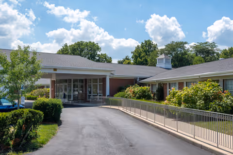 Front entrance of a single-story senior living building with a covered drop-off, wheelchair ramp, and landscaped beds under a blue sky.