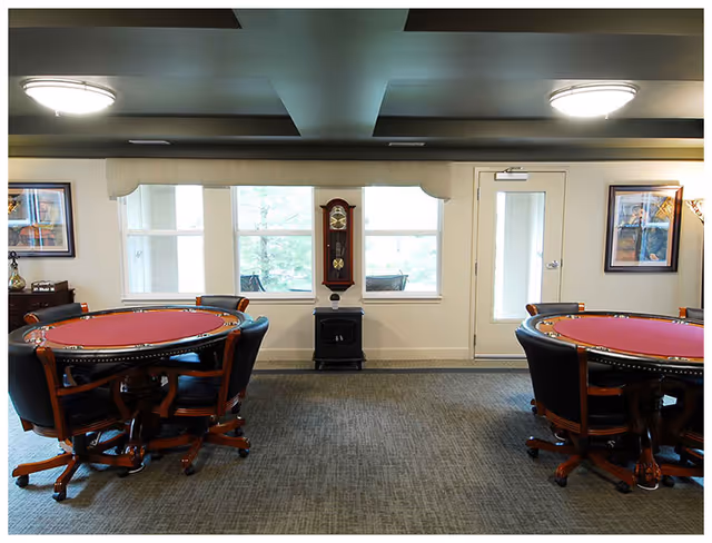Interior room with two round poker tables covered in red felt, each surrounded by black leather chairs with wooden legs. The room has a gray carpet, three large windows with a wall clock between them, a glass door, and framed artwork on the walls. Ceiling lights provide illumination.