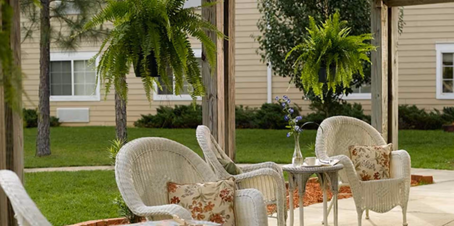 Wicker patio chairs and a small table under a pergola in a grassy courtyard with hanging ferns and a beige building in the background.
