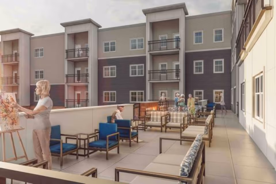 Outdoor patio area at Traditions of Mill Creek with several people engaging in activities. One person is painting on an easel, while others are seated or standing near a fire pit surrounded by cushioned chairs and sofas. The patio is adjacent to a multi-story building with balconies and windows.