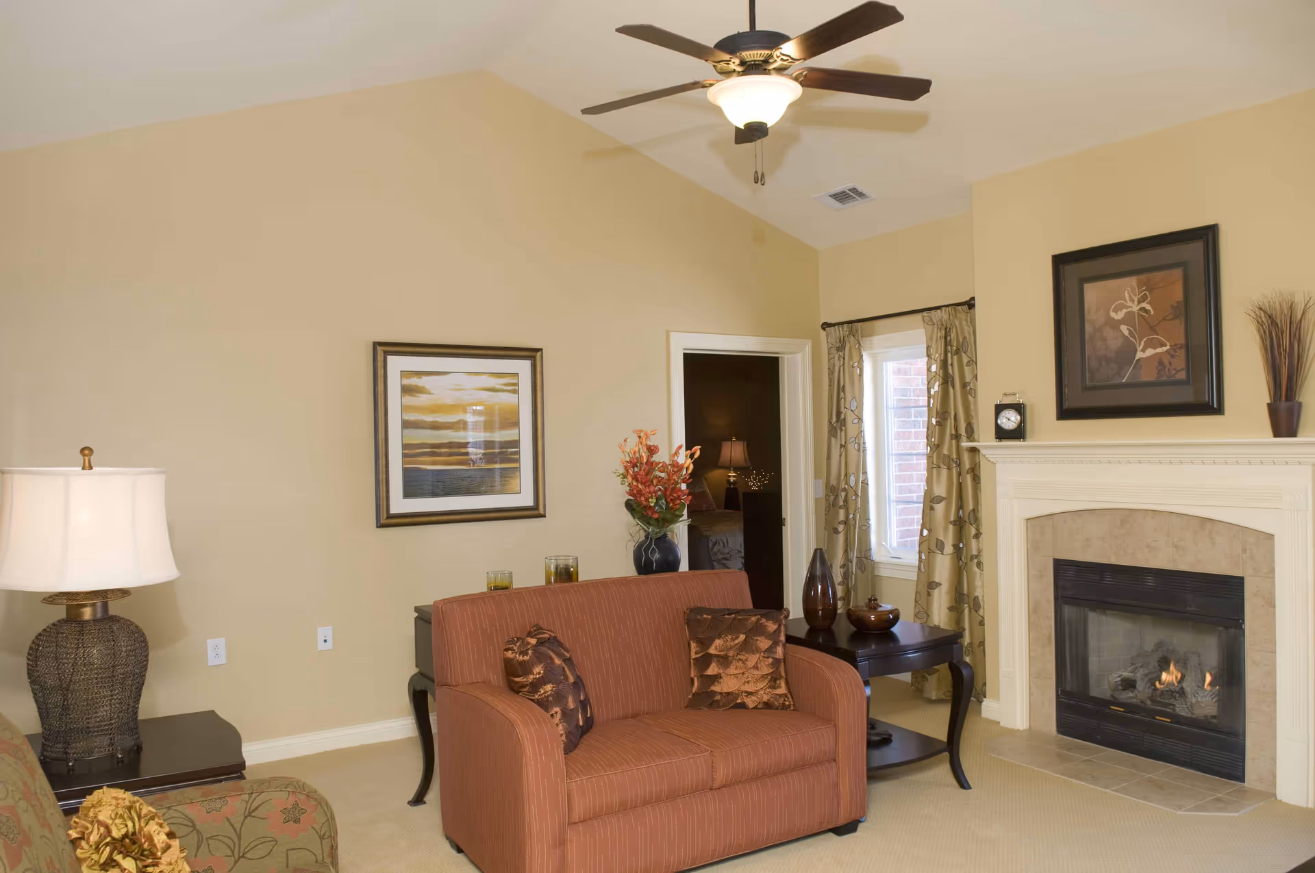 A cozy living room with beige walls and carpet, featuring a red loveseat with two decorative pillows, a fireplace with a lit fire, a side table with vases, a lamp on a dark wooden table, framed artwork on the walls, and a ceiling fan with a light fixture.