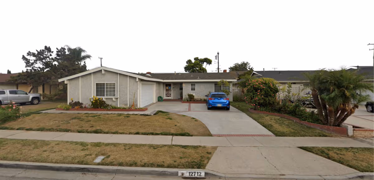 Single-story residential building with a driveway and a blue car parked in front. The house has a light-colored exterior with a garage on the left side and a front door in the center. There are some bushes and small trees in the front yard, and a sidewalk runs along the street in front of the house.