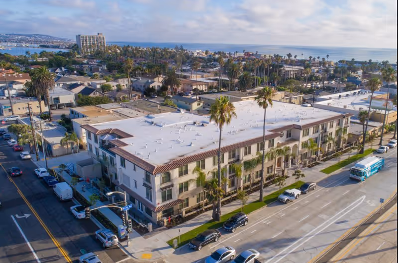 Aerial view of a three-story Mediterranean-style senior living building by the coast with palm trees and surrounding streets.