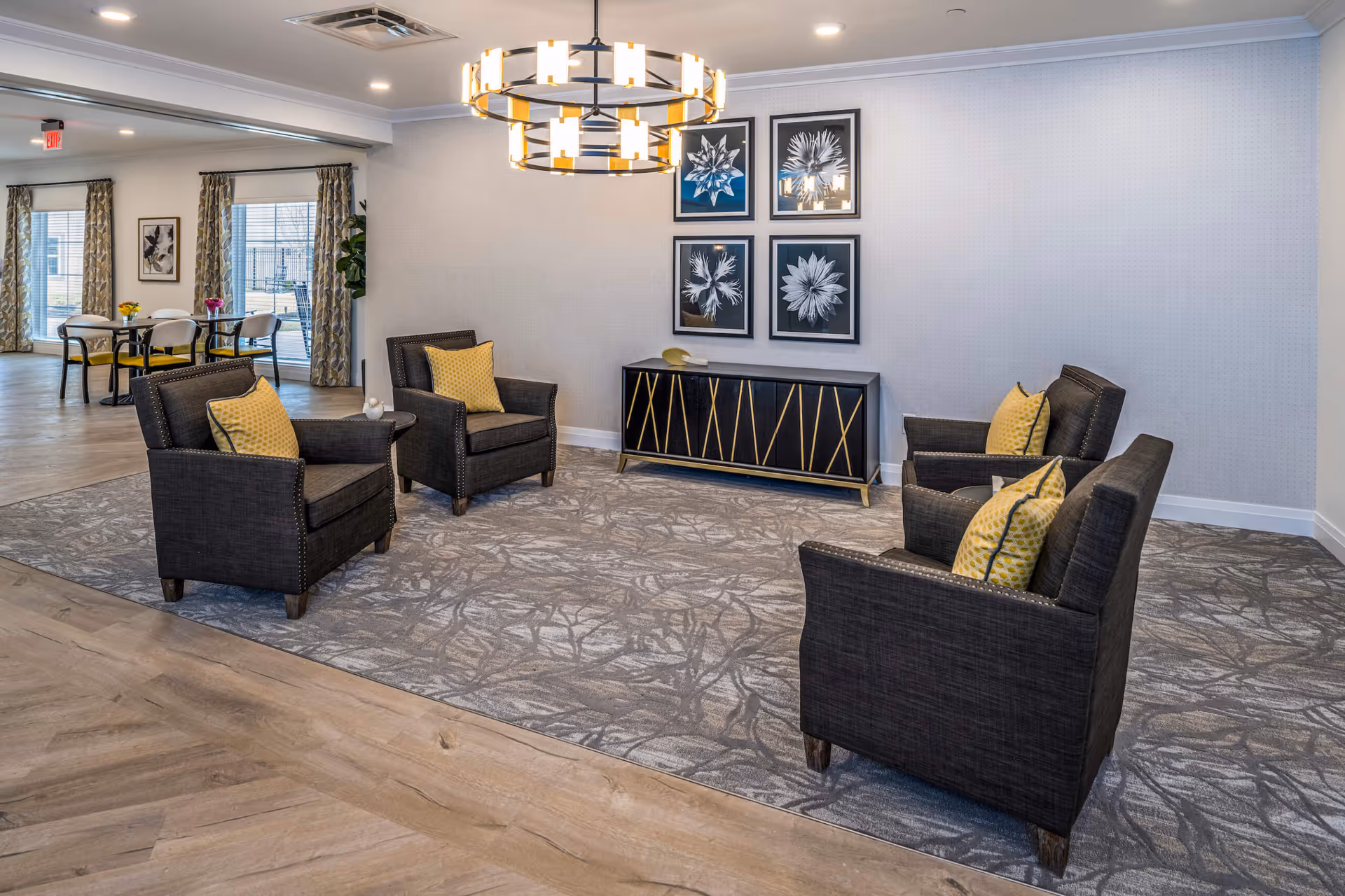 Stylish communal lounge with four dark armchairs with yellow pillows arranged on a patterned rug, a decorative sideboard and framed artwork on the wall, with dining tables visible in the background.