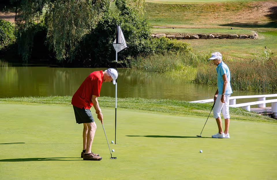 Two elderly people playing golf on a putting green near a small pond with greenery and trees in the background. One person is putting while the other watches.