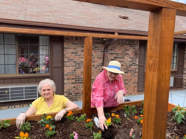 Two elderly women gardening in a raised wooden planter bed outside a brick building. One woman is wearing a yellow shirt and gloves, while the other is wearing a pink shirt and a wide-brimmed hat. There are orange and yellow flowers planted in the soil.