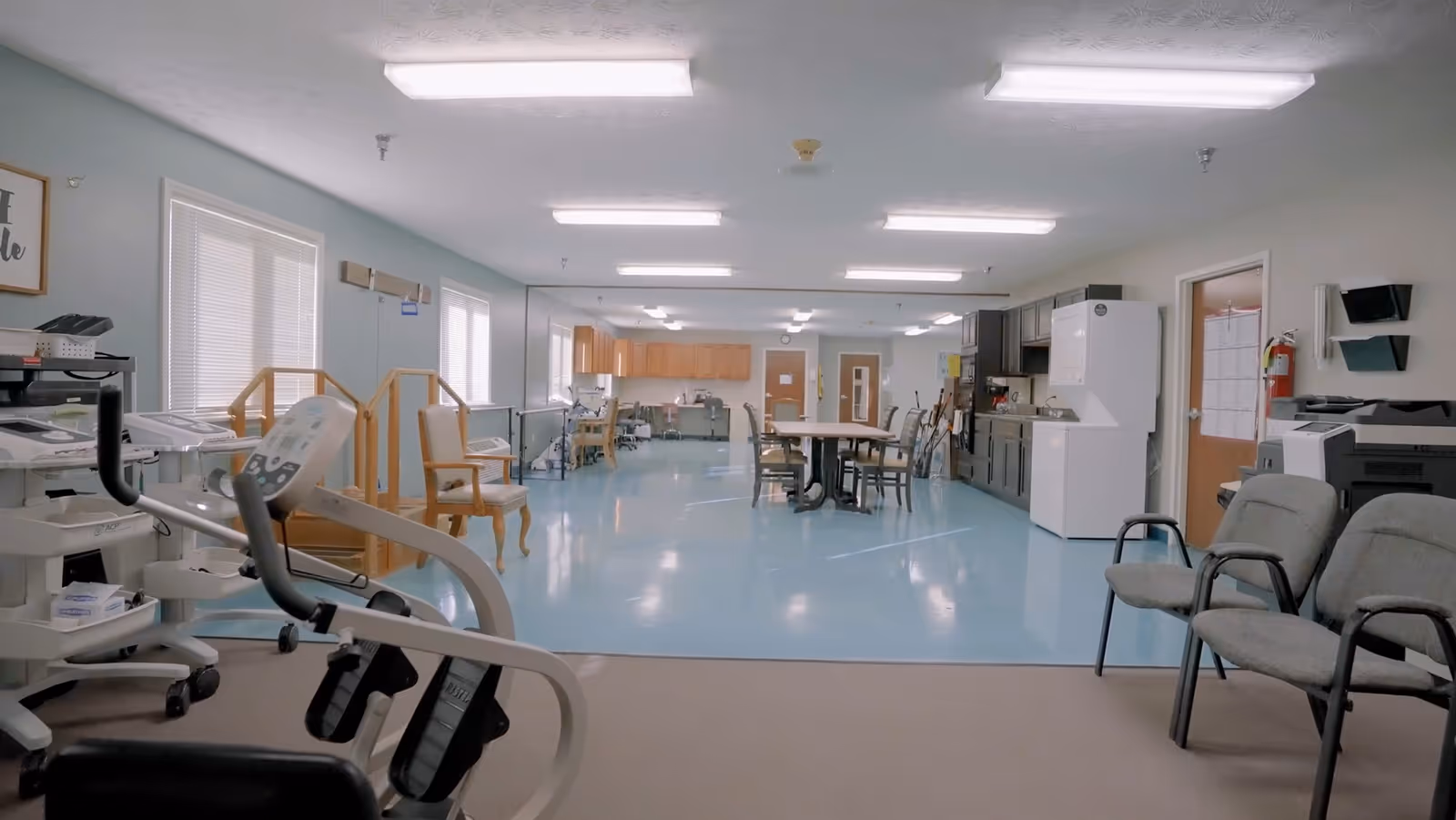 A spacious rehabilitation and healthcare center room with exercise equipment on the left, chairs along the walls, a table with chairs in the center, and a kitchenette area with cabinets, a refrigerator, and a microwave on the right. The room has bright overhead lighting and large windows letting in natural light.