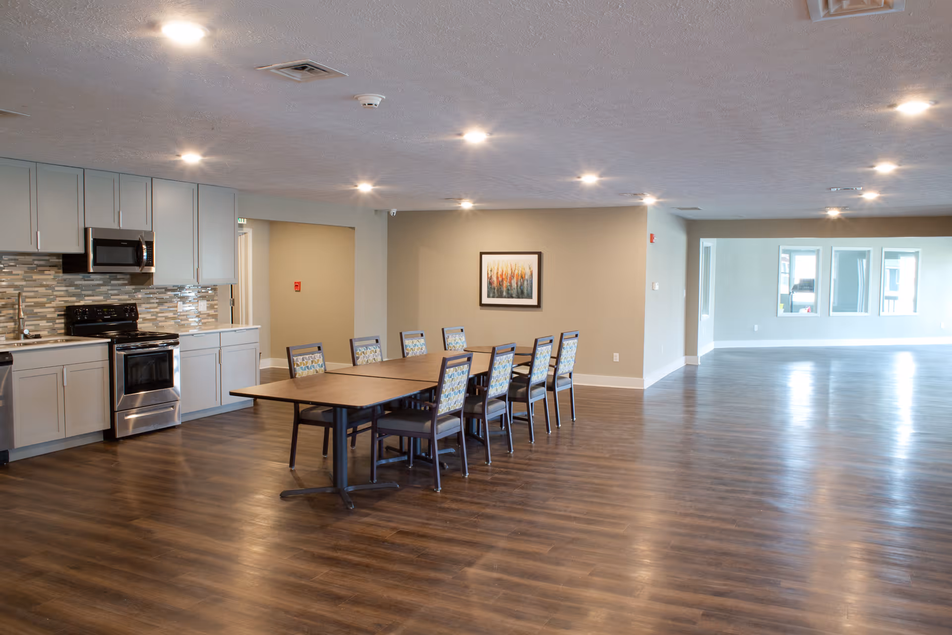 A spacious interior room featuring a modern kitchen area with gray cabinets, a stainless steel stove and microwave, and a tiled backsplash. In front of the kitchen is a long table with eight chairs. The room has wood flooring, recessed ceiling lights, and a framed artwork on the wall. Large windows are visible in the background, allowing natural light into the space.