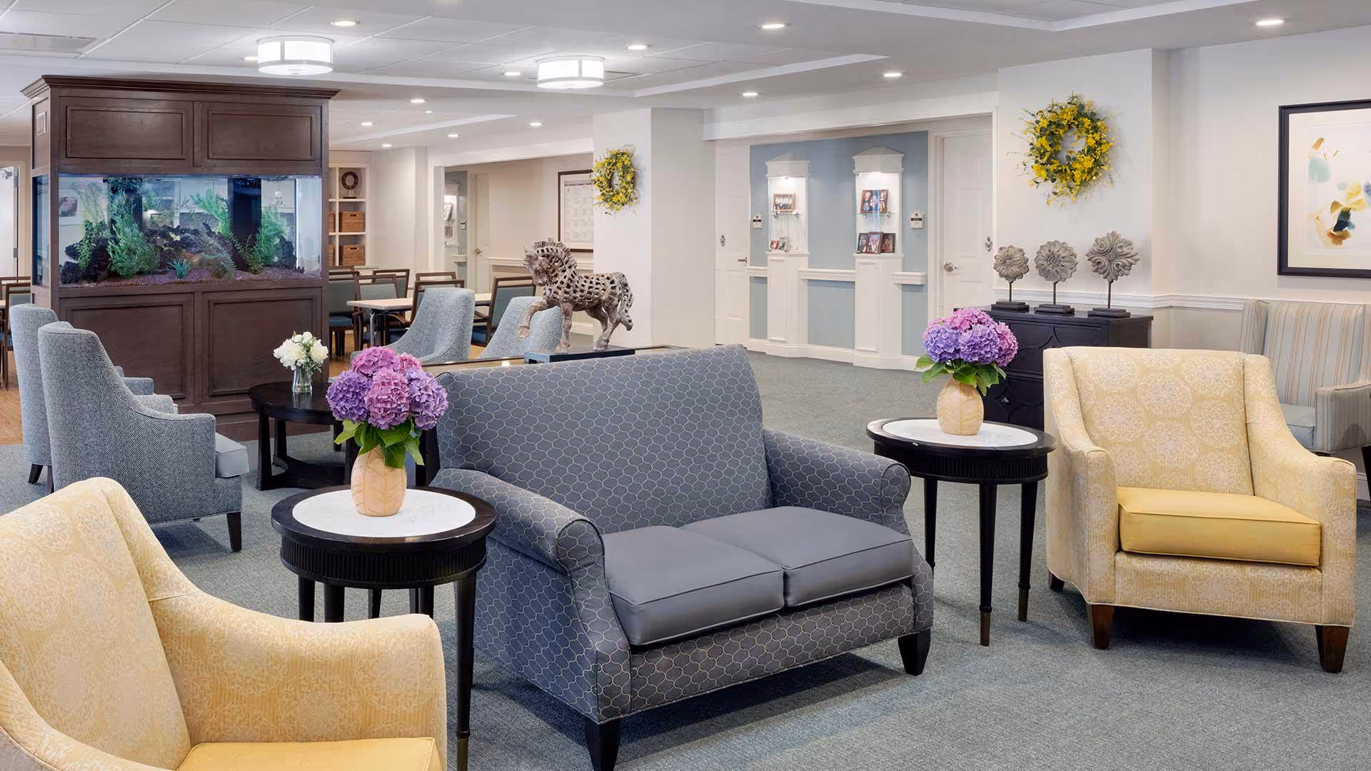 A well-lit senior living facility common area with comfortable seating including a gray patterned loveseat and two yellow armchairs. There are small round tables with vases of purple flowers beside the chairs. In the background, there is a large wooden aquarium, additional seating, decorative wall art, and a sculpture of a horse. The room has a calm and inviting atmosphere with soft lighting and neutral-colored walls.