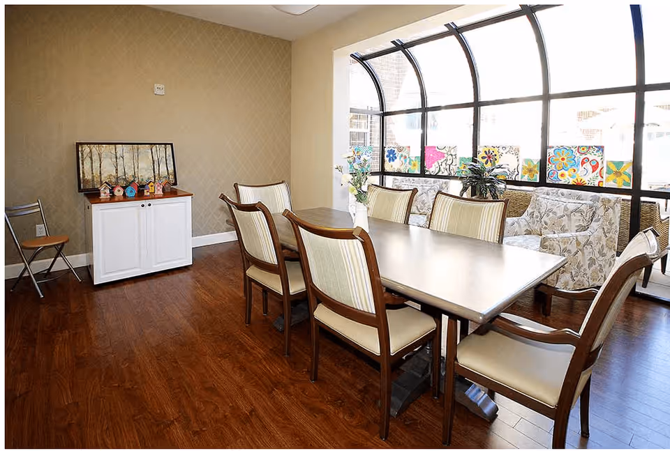 A dining area with a rectangular table surrounded by six cushioned chairs. The room has wooden flooring and a large curved window with colorful flower paintings on the lower part. There is a white cabinet with small decorative birdhouses and a framed picture on top, and a folding chair is placed against the wall.