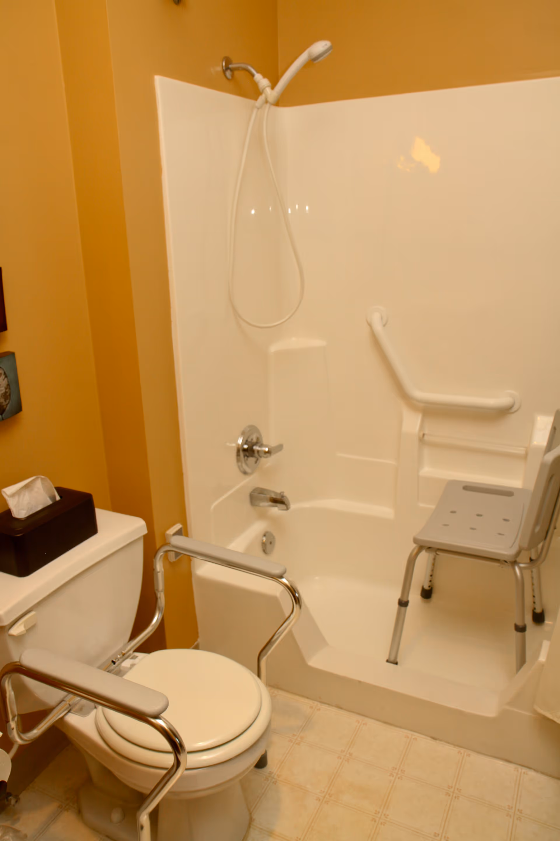 A bathroom with a white toilet equipped with metal safety rails and a tissue box on top. Next to the toilet is a white bathtub with a handheld showerhead, a grab bar, and a gray shower chair inside the tub. The walls are painted a warm yellow color.
