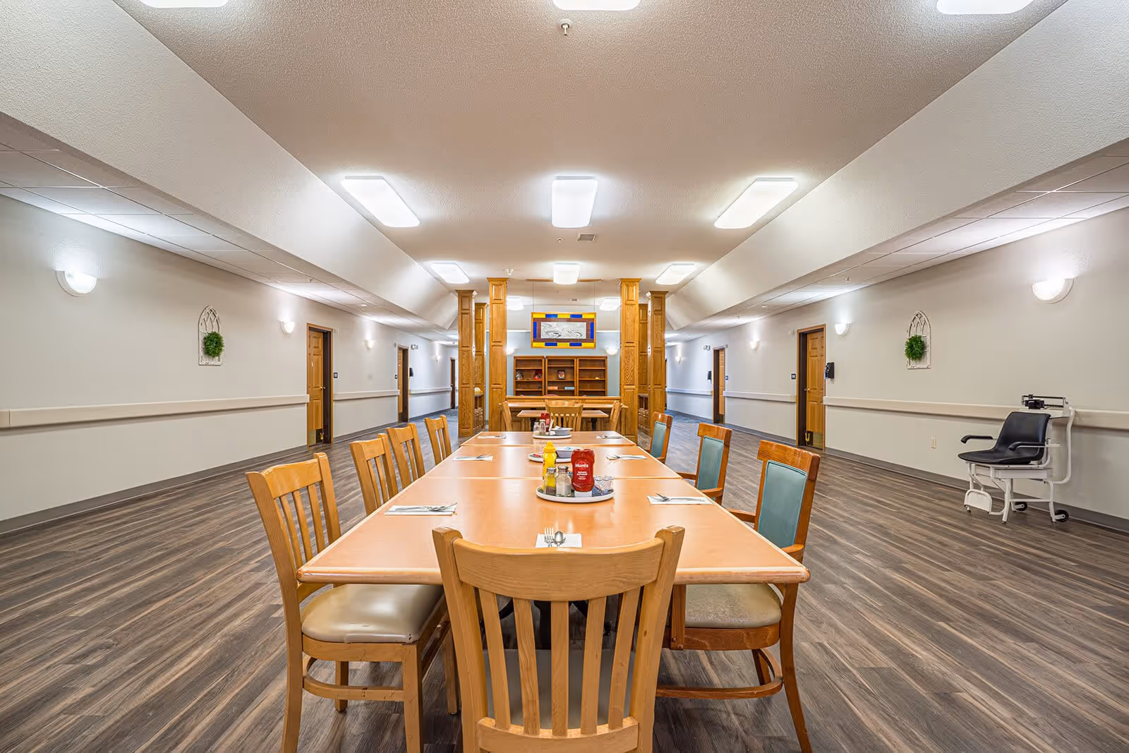 A long dining table set with chairs in a spacious room with wood flooring and white walls. The table has condiments and utensils placed on it. The room has multiple doors along the walls, bright ceiling lights, and decorative wall hangings. There is a black chair with wheels on the right side of the room.
