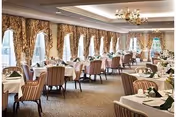 Elegant dining room with multiple tables covered in white tablecloths, each set with folded napkins and silverware. The room features large windows with ornate drapes, striped upholstered chairs, chandeliers, and carpeted flooring.