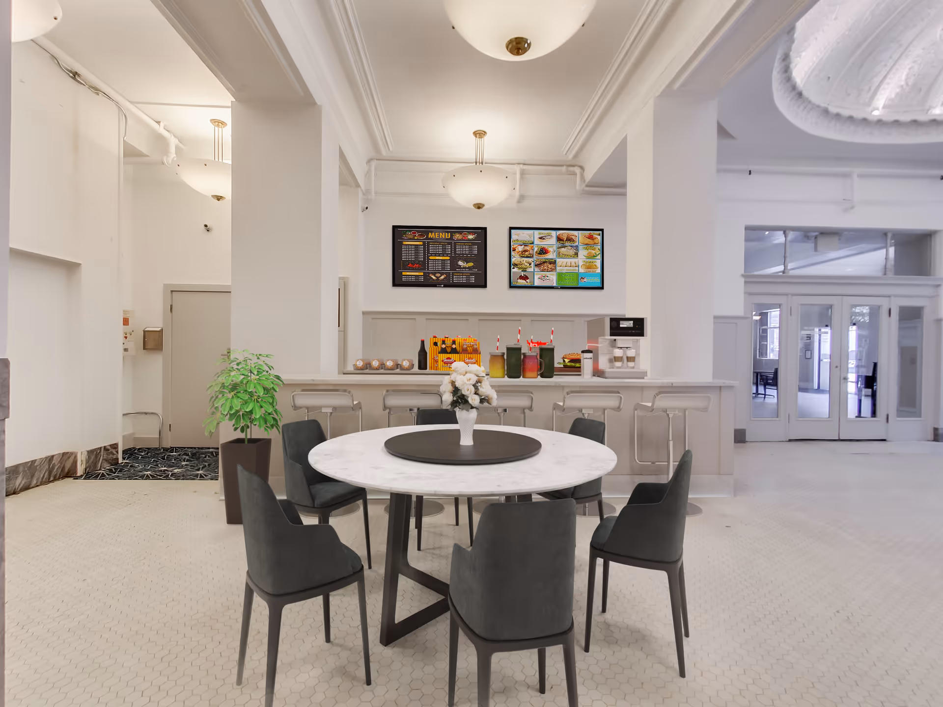 A bright dining area with a round white marble table surrounded by six dark gray chairs. On the table is a white vase with white flowers. Behind the table is a counter with several bar stools, drinks, and snacks displayed. Two menu boards are mounted on the wall above the counter. The room has white walls, a tiled floor, and large glass doors leading to another area.