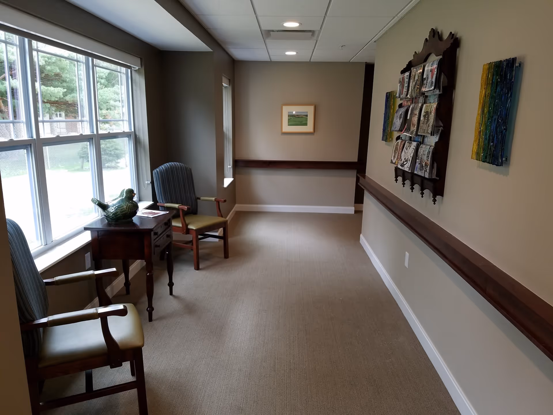 A quiet interior corridor with chairs and a small table by a large window, and a magazine rack and artwork on the wall.