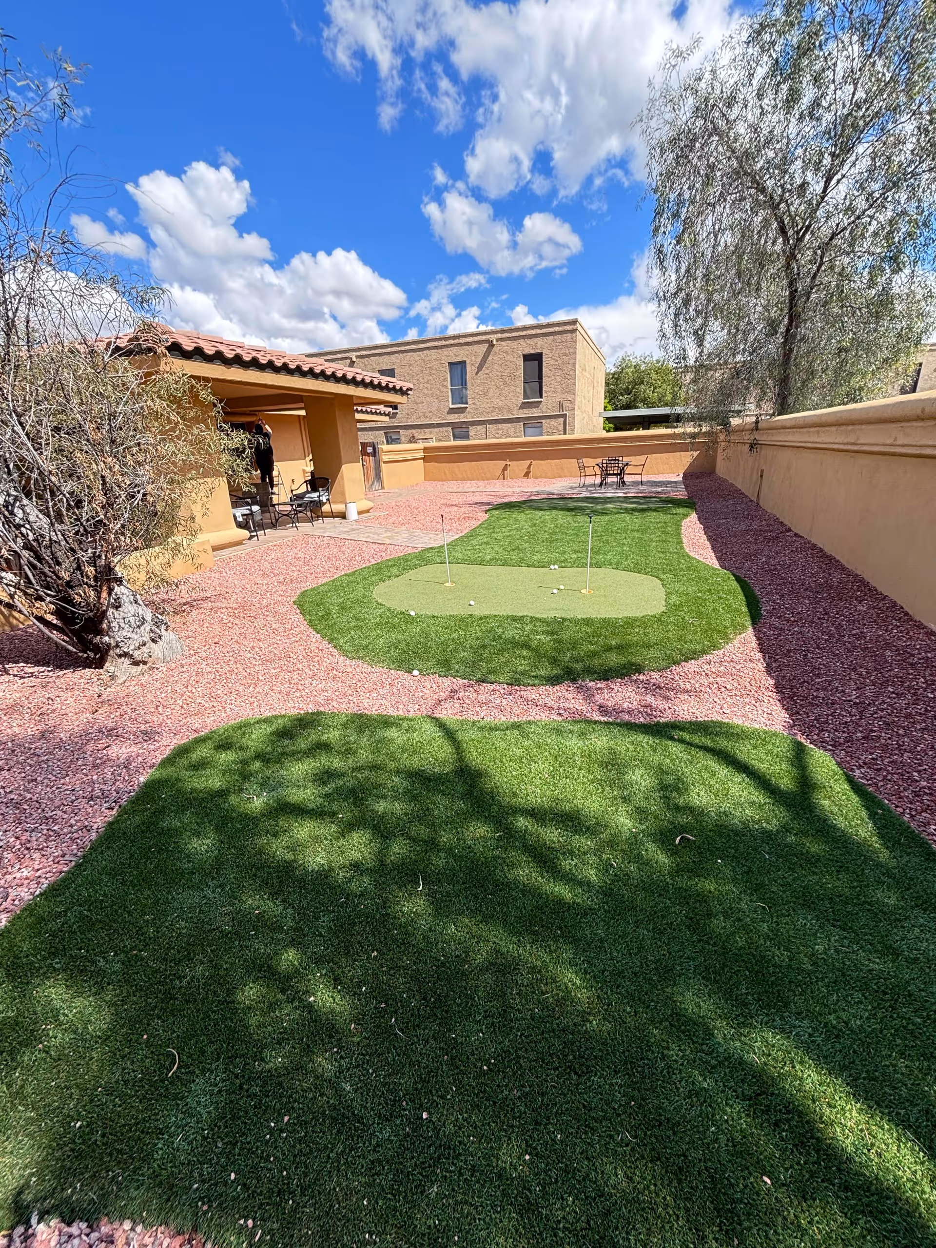 Outdoor area at Assisted Living of Mesa featuring a small putting green with three golf holes, surrounded by reddish gravel and a tan stucco wall. There are chairs and a shaded patio area with a tiled roof on the left side, and a tree casting shadows on the green grass. The sky is blue with scattered clouds.
