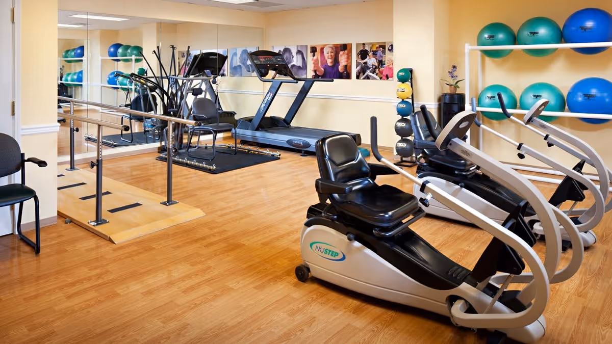 A senior living facility exercise room with various fitness equipment including recumbent exercise bikes, a treadmill, an elliptical machine, and a set of parallel bars. The room has wooden flooring, a wall-mounted rack with large exercise balls, and framed photos of seniors on the wall.