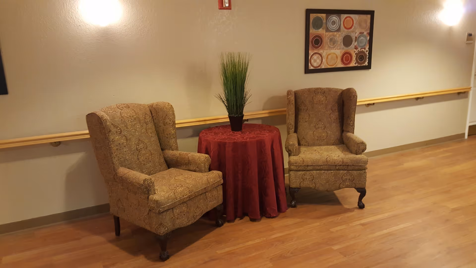 Two patterned wingback chairs flanking a small round table draped in red with a potted plant in a hallway with hardwood floors and wall art.