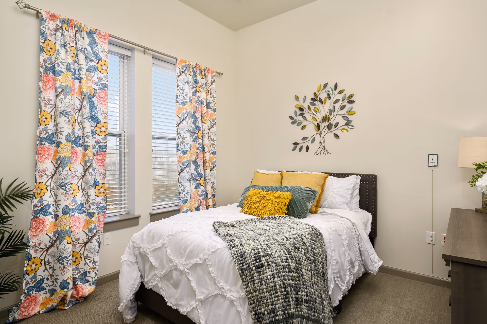 A cozy bedroom with a bed dressed in white bedding, accented with yellow and green pillows and a textured throw blanket. The room features floral patterned curtains on two windows, a decorative metal tree wall art above the bed, a bedside lamp, and a small plant on a wooden dresser.