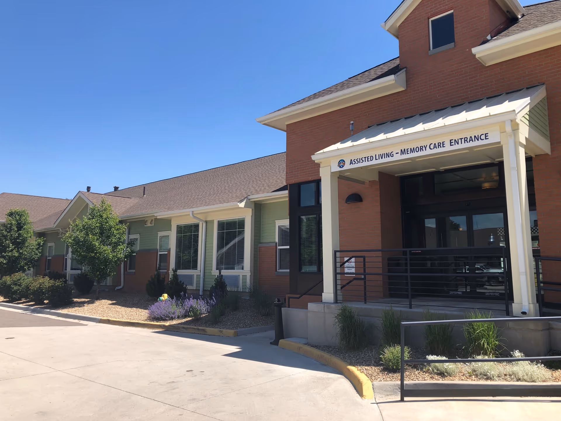 Exterior view of the Mountain Vista Assisted Living & Memory Care facility showing the entrance with a sign that reads 'Assisted Living - Memory Care Entrance'. The building has a combination of brick and green siding with several windows, landscaping with bushes and flowers, and a clear blue sky above.