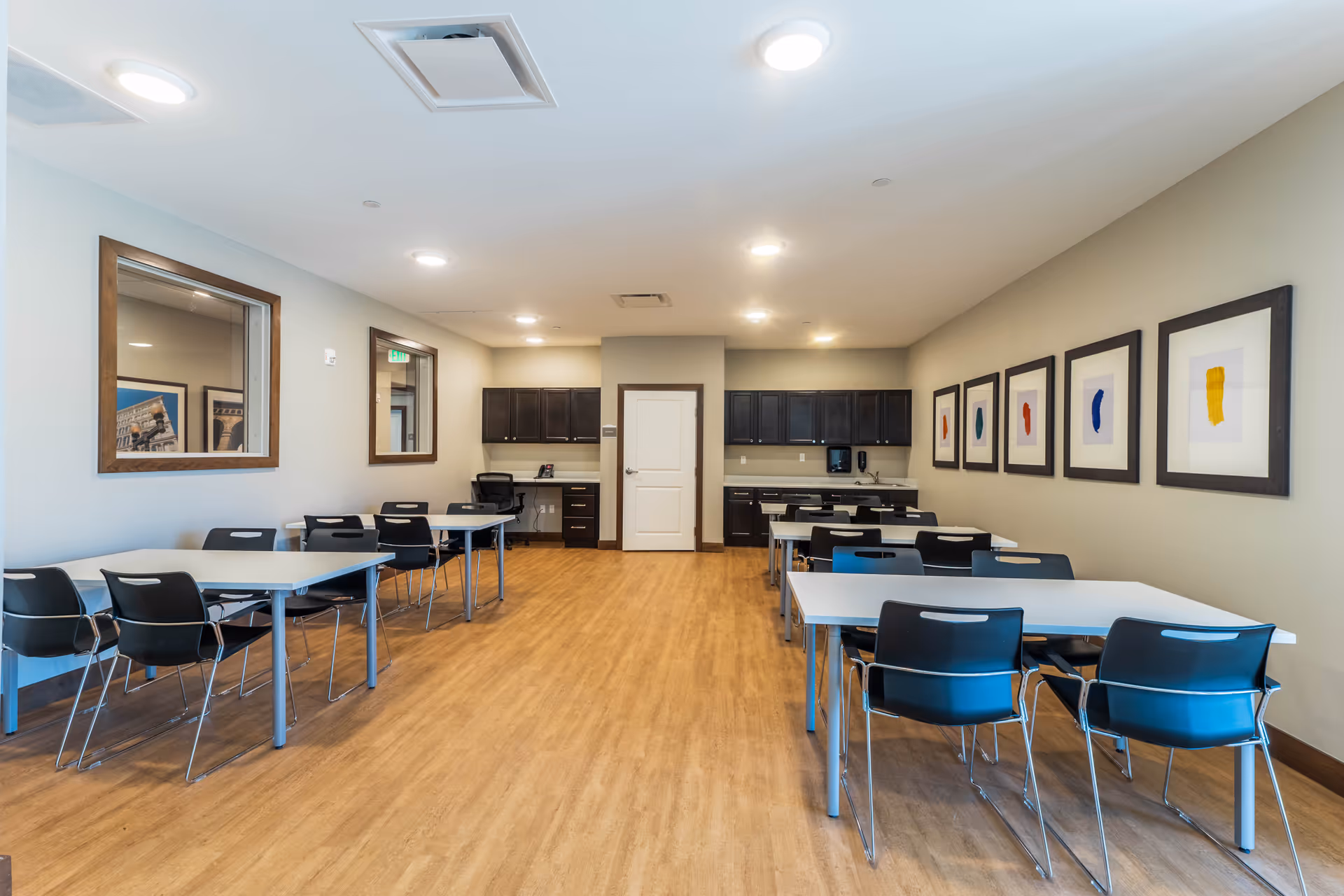 A clean and modern dining room with several tables and black chairs arranged neatly. The room has light-colored walls adorned with framed abstract art, wood flooring, and a small kitchenette area with dark cabinets and a countertop at the far end.