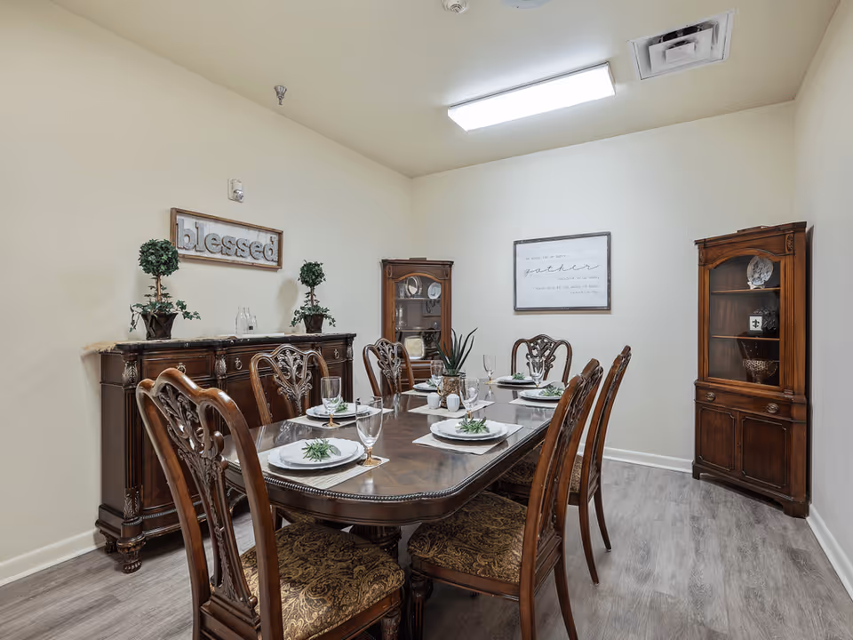 A formal dining room with a wooden dining table set for six people. The table has white plates, glasses, and decorative greenery on each place setting. The room features two wooden display cabinets and a wooden sideboard with two potted plants and decorative items. The walls are light-colored with framed signs that read 'blessed' and 'gather'. The floor is a light wood laminate.
