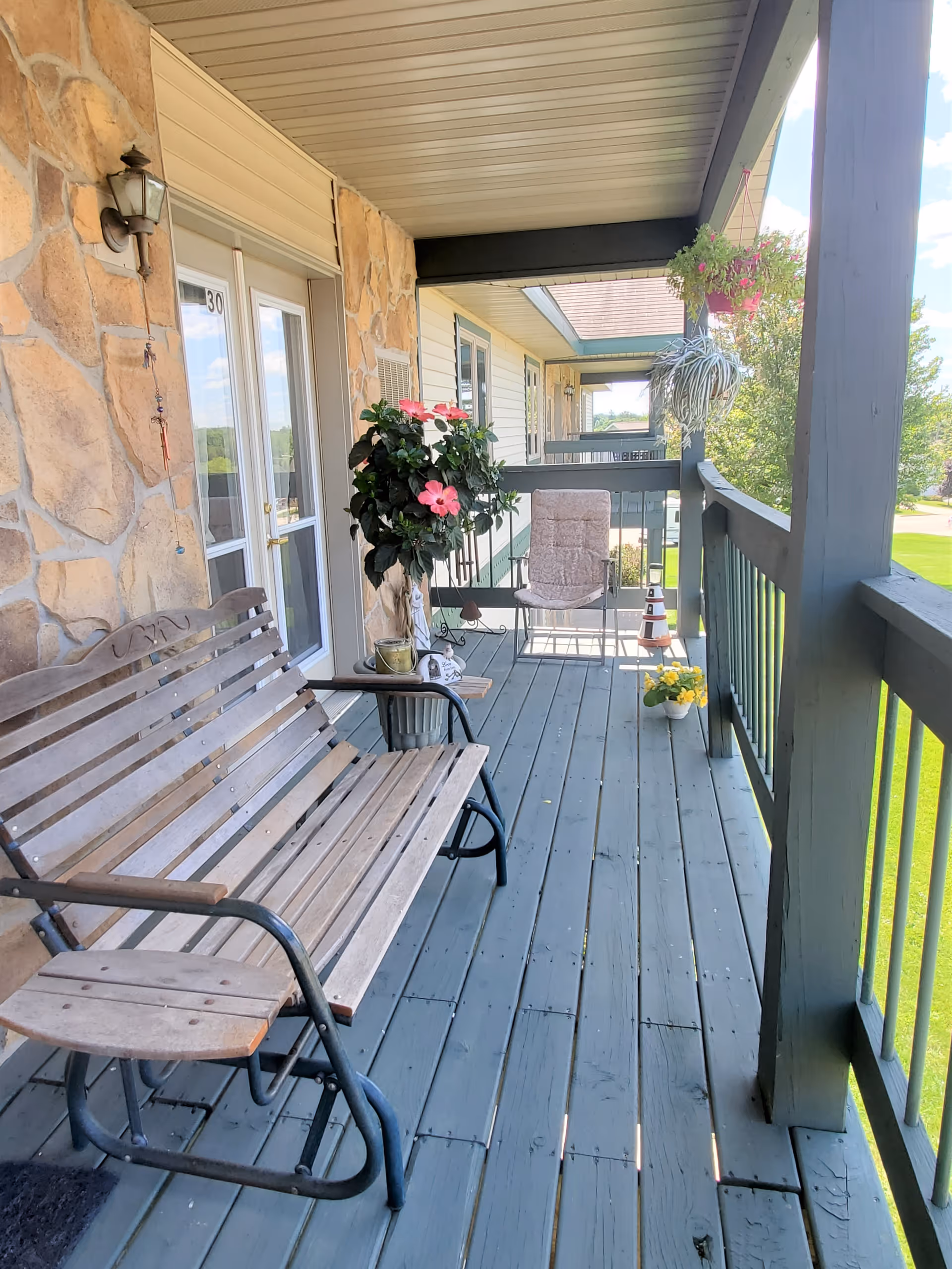 A covered outdoor balcony with a wooden bench swing, a cushioned chair, and several potted plants including a large flowering plant and hanging greenery. The balcony has a stone wall on one side and overlooks a green lawn with trees in the background.
