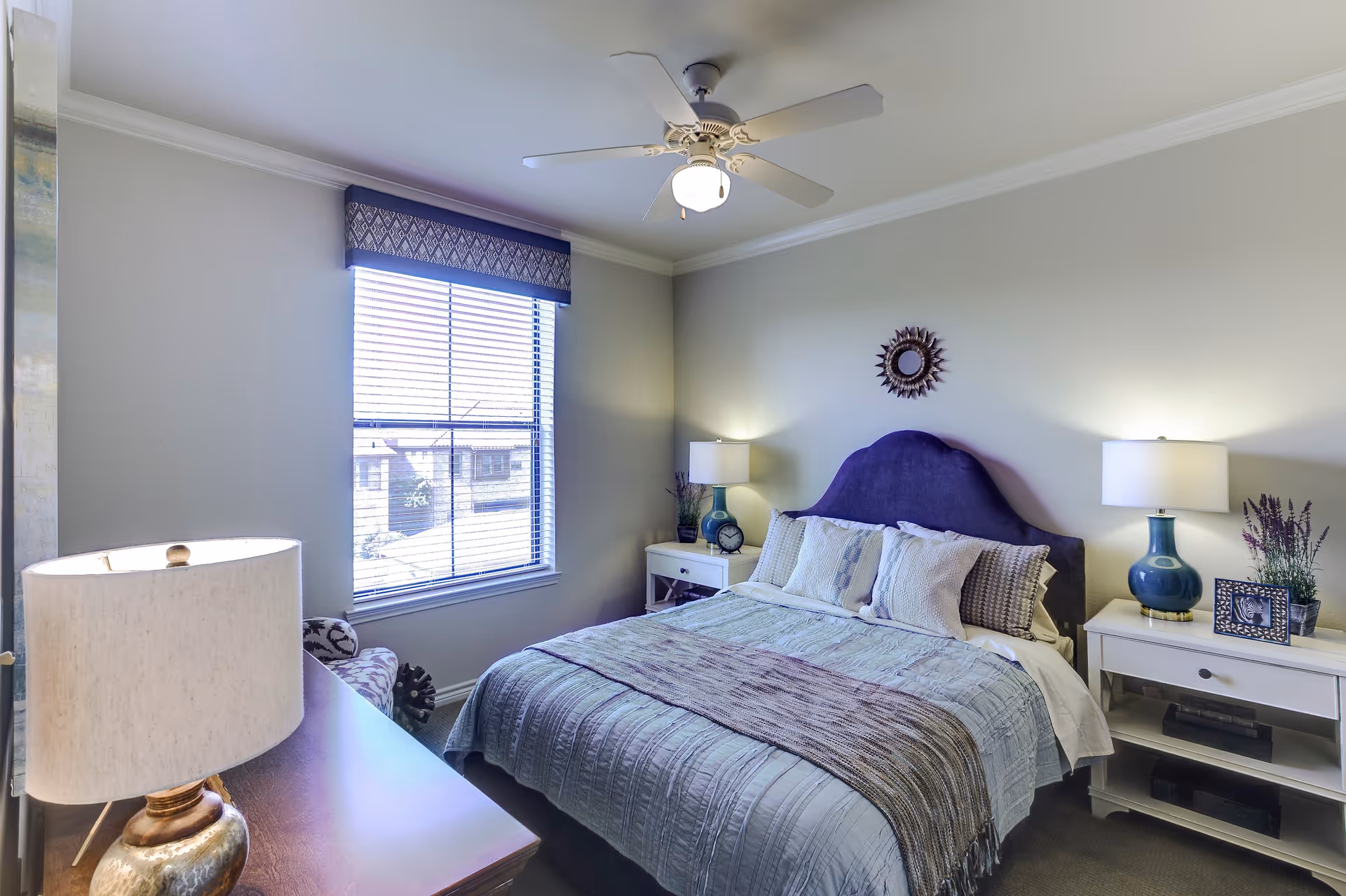 A well-lit bedroom with a large bed featuring a purple headboard, multiple pillows, and a light blue bedspread with a brown throw blanket. There are two white nightstands on either side of the bed, each with a blue lamp and decorative items including a clock, a framed photo, and a plant. A window with blinds and a patterned valance lets in natural light. A ceiling fan with a light fixture is mounted on the ceiling.