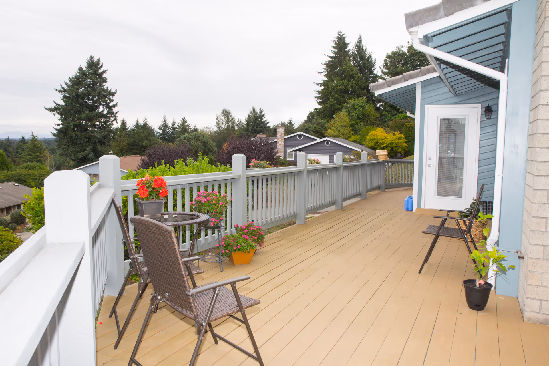 A spacious outdoor deck with light brown wooden flooring and a gray railing. The deck has several potted plants with colorful flowers and two brown wicker chairs around a small glass-top table. The deck is attached to a blue house with a white door and a small overhang. Trees and neighboring houses are visible in the background under a cloudy sky.