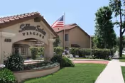 Entrance sign for Soldano Senior Village Apartments with an American flag flying above. The sign is surrounded by well-maintained landscaping including green grass, bushes, and trees. A sidewalk runs alongside the entrance, and a two-story building is visible in the background under a clear sky.