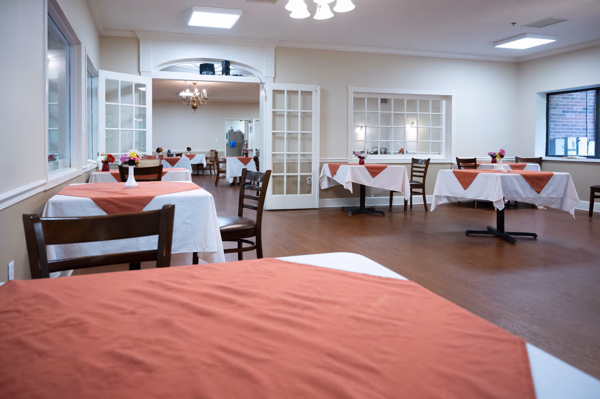 Dining room with multiple tables covered with white tablecloths and rust-colored table runners. Each table has a small vase with flowers. Wooden chairs are arranged around the tables. The room has light-colored walls, wooden flooring, and a window showing an exterior brick wall. French doors lead to another room with additional tables and a chandelier.
