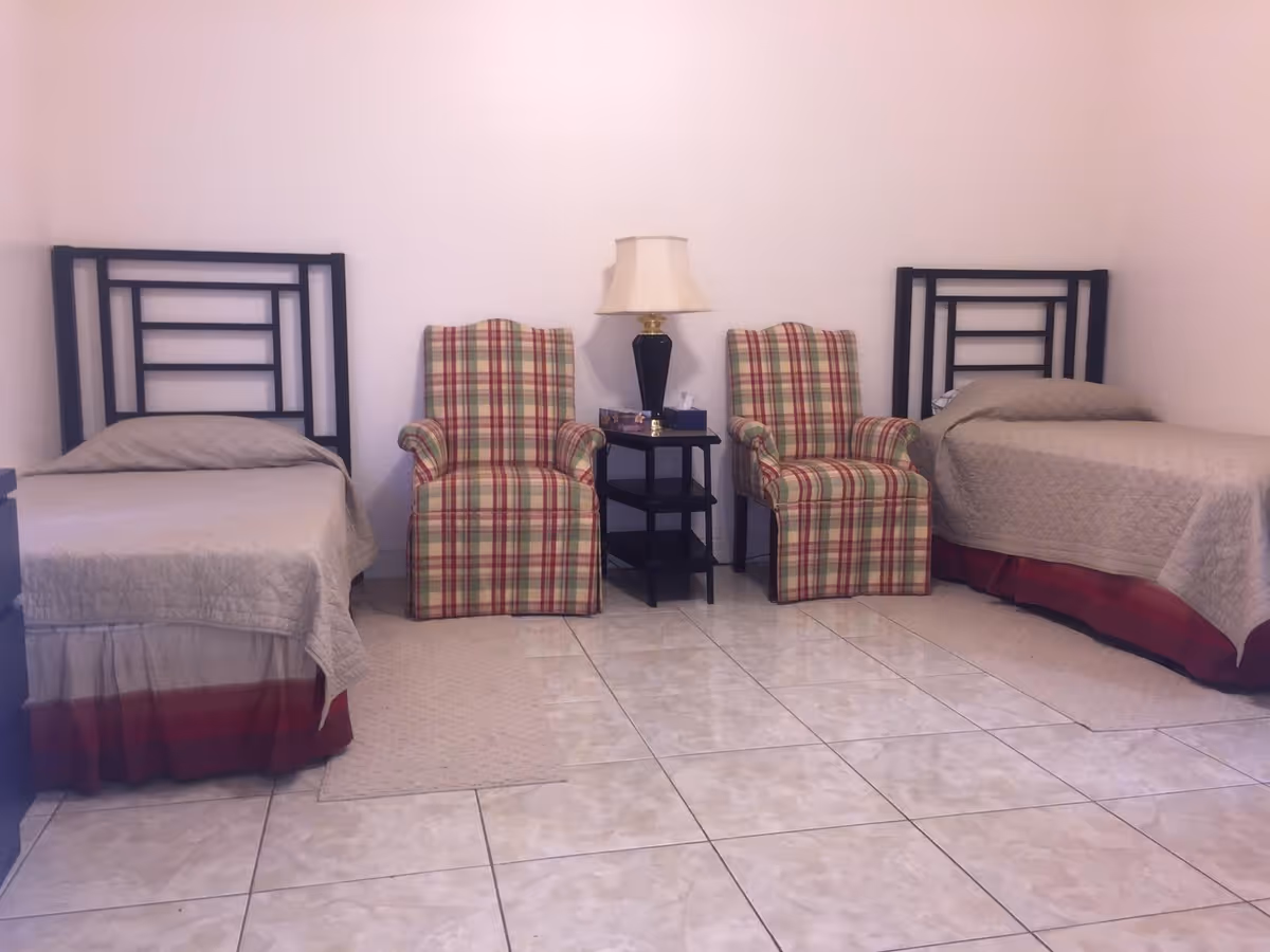 A simple bedroom with two single beds, each with a beige quilt and red bed skirt. Between the beds are two plaid upholstered armchairs with red, beige, and green stripes, and a small black side table with a lamp on it. The floor is tiled with light-colored tiles, and the walls are plain white.