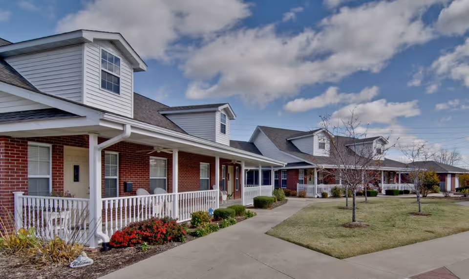 Exterior front view of a single-story brick senior living building with covered porches, white railings, dormer windows, walkways and landscaped lawn.